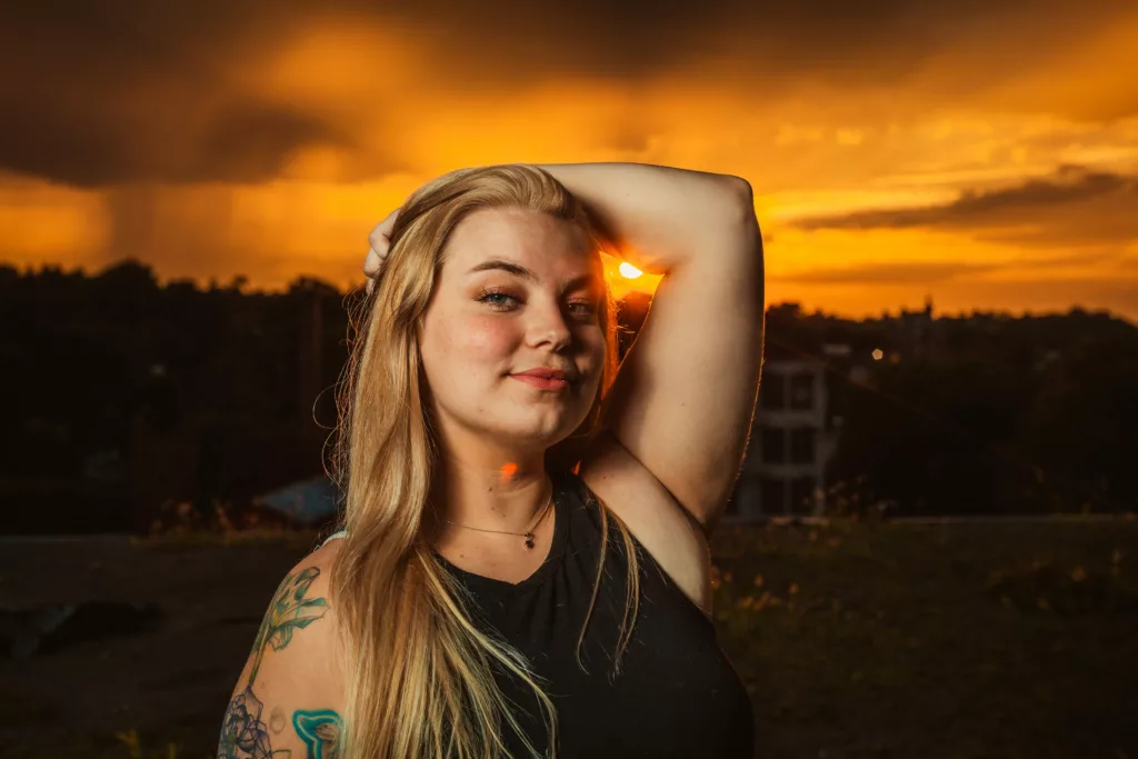 A person with long hair and a bright smile poses for a portrait against a sunset sky filled with clouds.