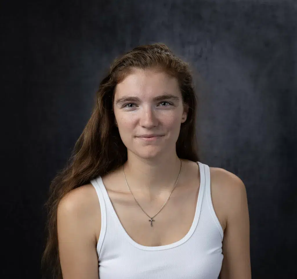 A smiling person in a sleeveless shirt and active tank is posing for a portrait, their chest and neck adorned with a necklace.