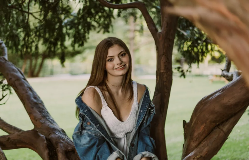 A person with long hair and a smiling face stands in a park, leaning against a tree trunk surrounded by grass.