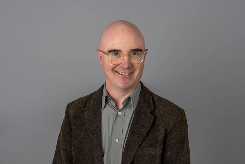 A gentleman wearing a dress shirt, blazer, and outerwear is smiling in a portrait headshot taken indoors.