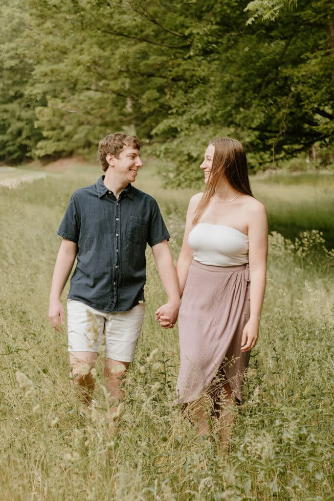 Two people are walking through a grassy field, holding hands and smiling at each other. They're surrounded by greenery and dressed in summer attire.