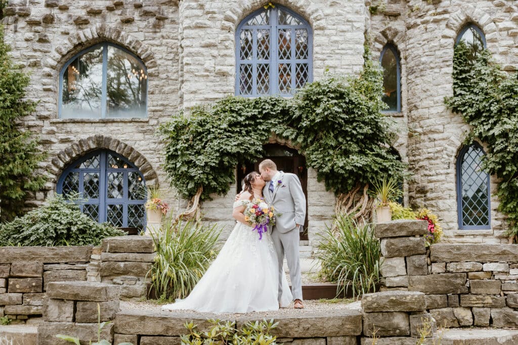 Two people in wedding attire kissing outside a stone building covered with ivy, surrounded by greenery and flowers, implying a wedding ceremony.