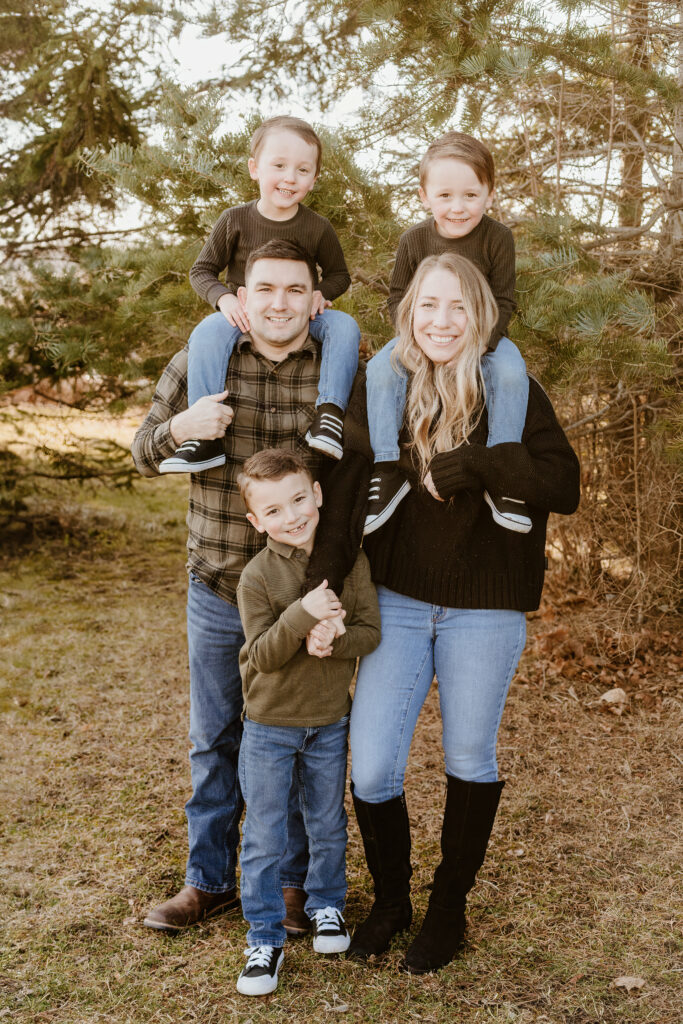 A family of five poses outdoors. Two persons with three children smile, surrounded by trees. Two kids sit on shoulders, the youngest stands in front.
