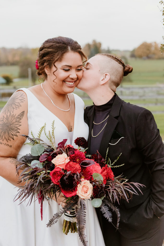 Two people dressed in wedding attire share a joyful moment outdoors. One person holds a bouquet of deep red and pink flowers, smiling, while the other kisses their cheek.