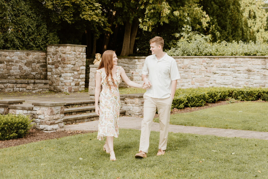 Two people are holding hands and walking in a lush garden with stone walls and steps. They appear happy and are casually dressed.