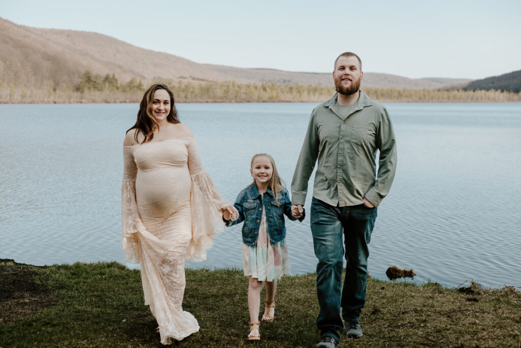 Two adults and a child walk hand-in-hand by a tranquil lake with forested hills in the background. They appear happy and connected in a natural setting.