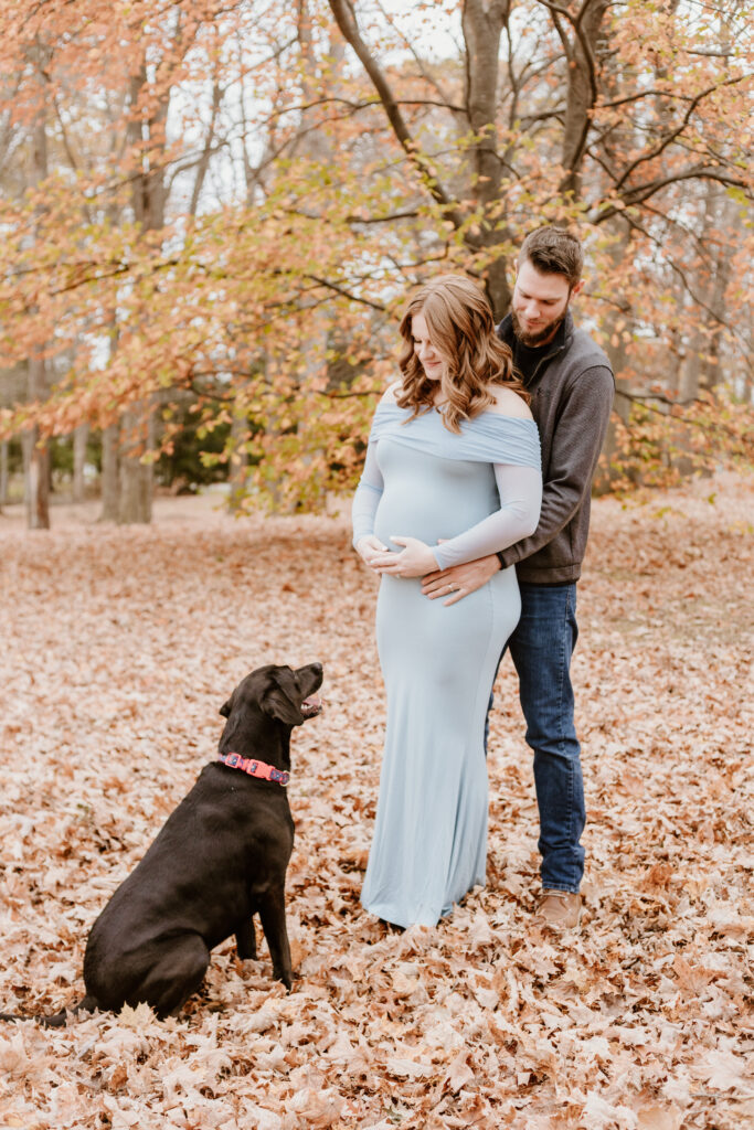 In an autumnal setting, a person embraces a pregnant person from behind while a black dog sits beside them, looking up attentively.