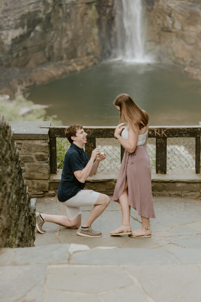 One person is kneeling in front of another, holding a small box, against a backdrop of a waterfall. Both look emotional in a rustic setting.