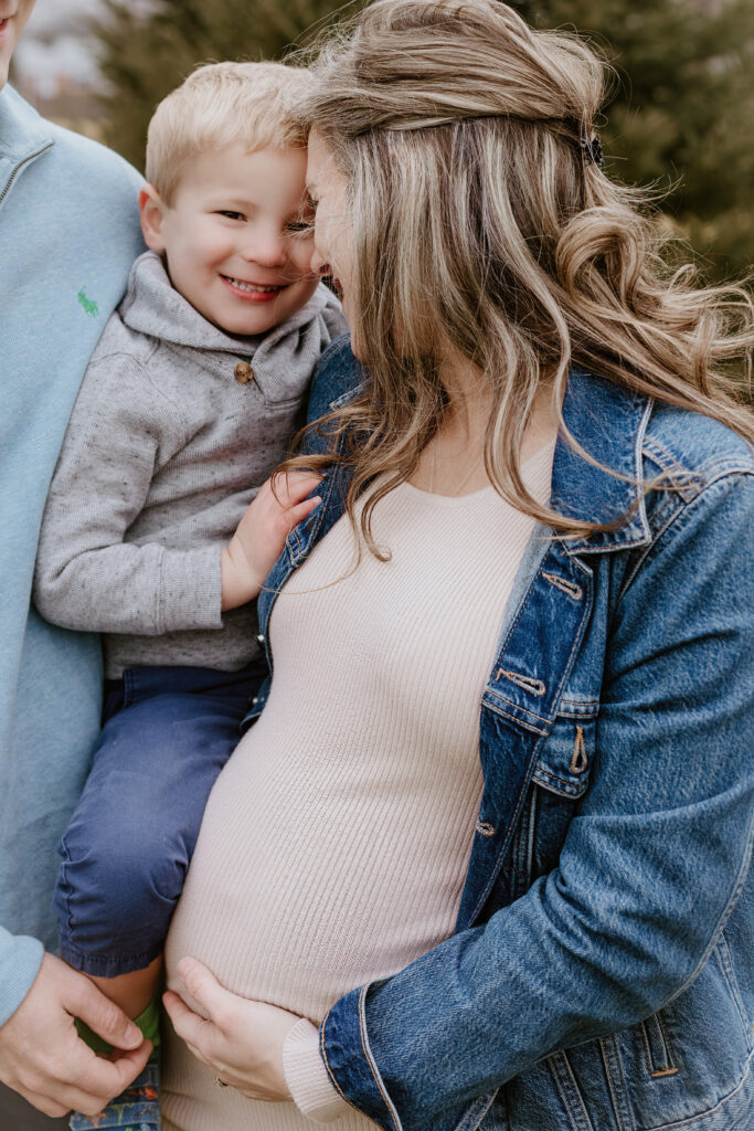 A joyful child with blonde hair smiles while holding the belly of a pregnant person wearing a denim jacket. Partial view of another adult present.