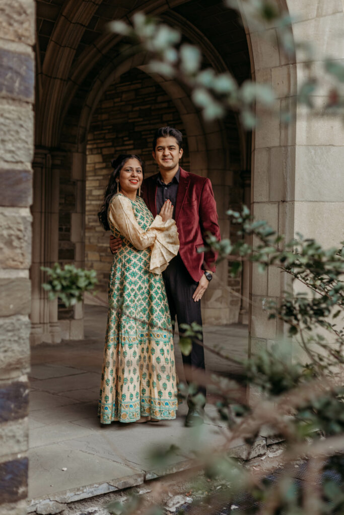 Two people stand under an archway, dressed in traditional Indian attire, surrounded by greenery, looking content and closely positioned to one another.