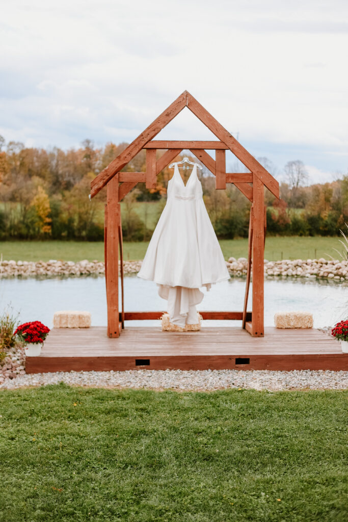 A wedding dress hangs on a rustic wooden archway by a pond, surrounded by greenery, flowering plants, hay bales, and autumnal trees in the background.