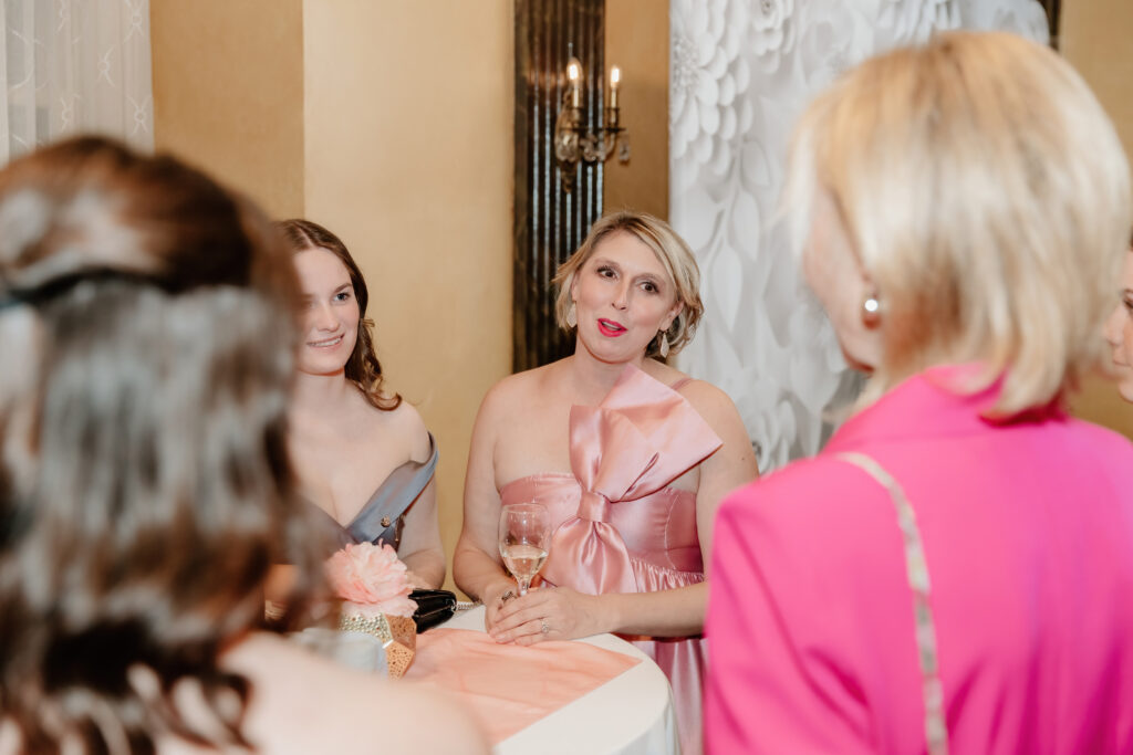Three people engaged in conversation at an indoor event. A person in a pink dress holds a wine glass, surrounded by elegant decor.