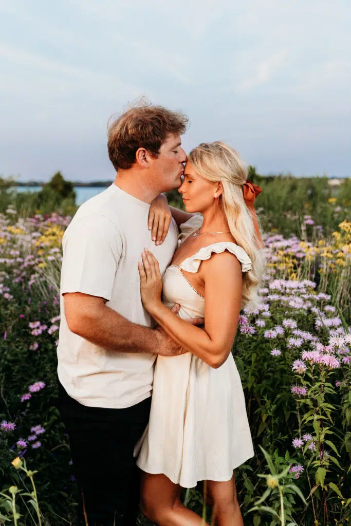 Two people embrace in a field of purple flowers. The person on the left kisses the forehead of the person on the right at sunset.