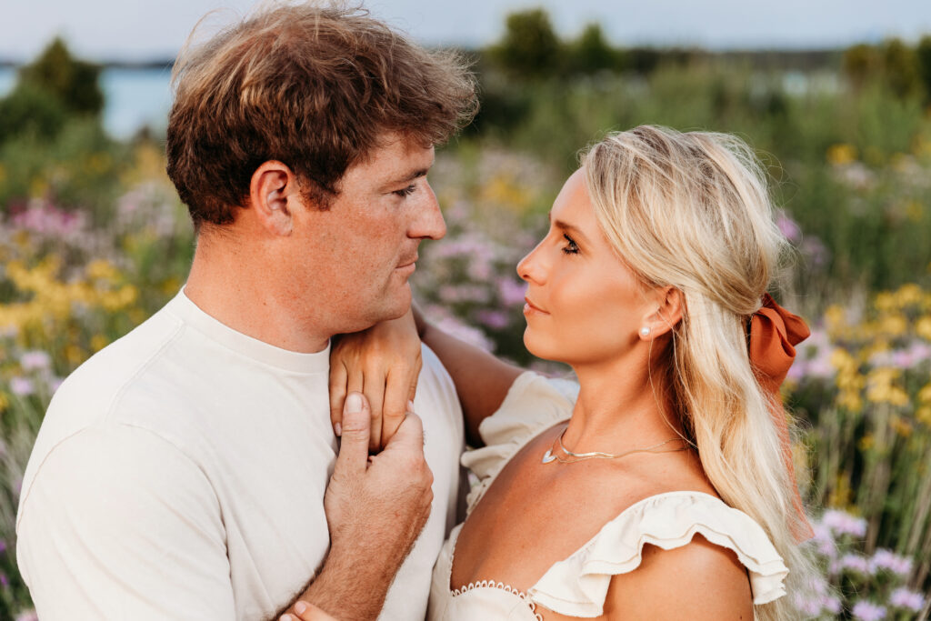 Two people are intimately facing each other in a field of wildflowers, with soft focus on a serene, natural background. They display a moment of connection.