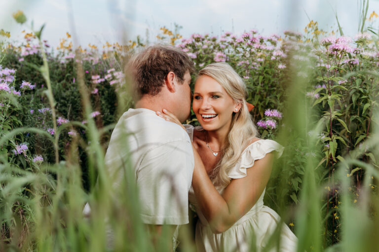 Two people are intimately engaging, surrounded by lush wildflowers. The person facing the camera smiles brightly, creating a cheerful, romantic atmosphere.