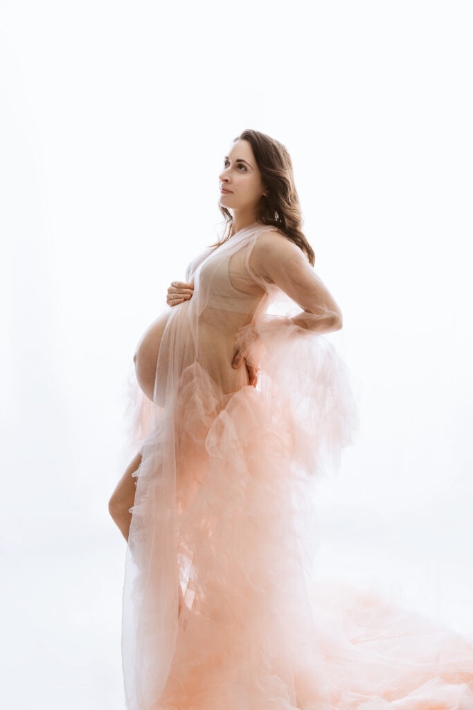 A person in a sheer, flowing, peach gown stands gracefully, cradling their abdomen, against a soft, white background.