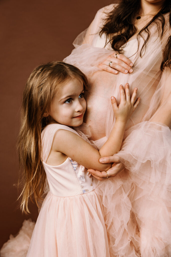 A child embraces a pregnant person, dressed in matching pink outfits, against a soft brown background, creating a tender and intimate moment.