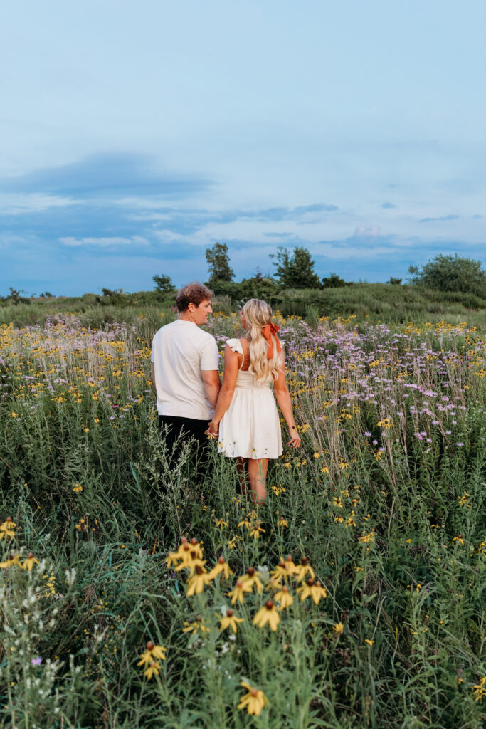 Two people are standing in a blooming field with purple and yellow flowers, looking into the distance under a cloudy sky. They appear relaxed and connected.