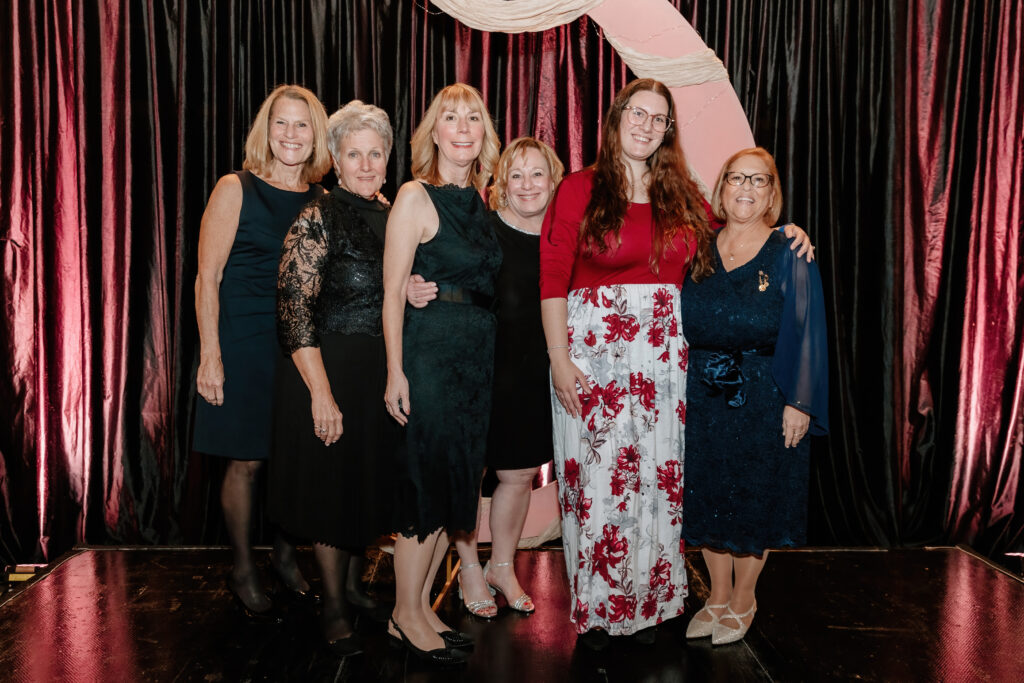 Six people posing together in front of a pink arch and dark curtains, dressed formally, standing on a shiny black stage surface.