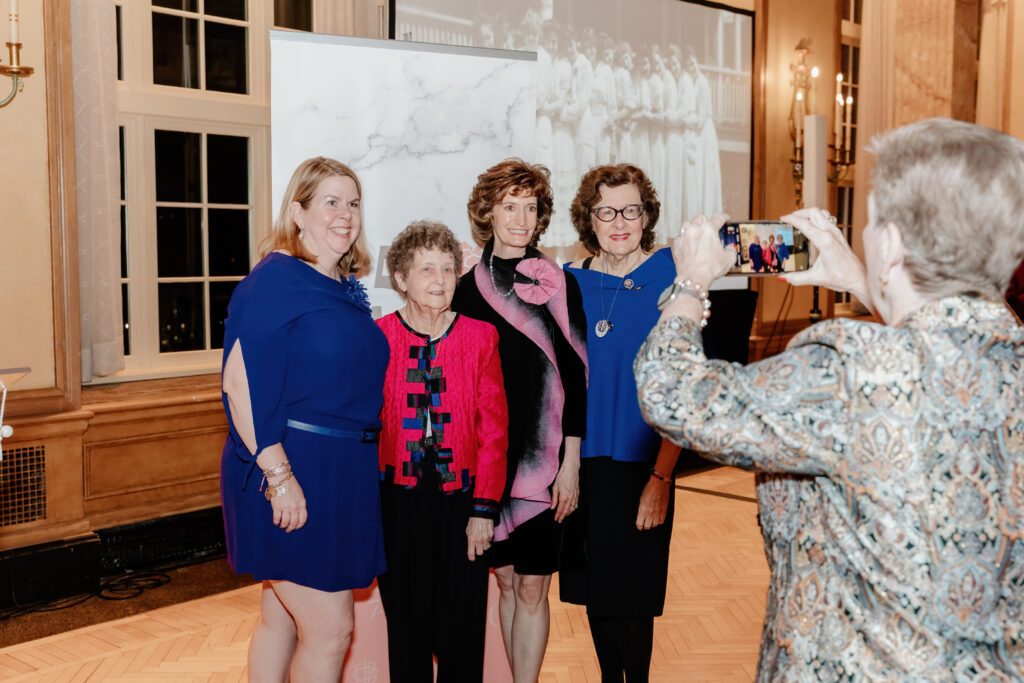 Four people pose for a photo indoors, one person takes a picture with a smartphone. A historical image is projected in the background.