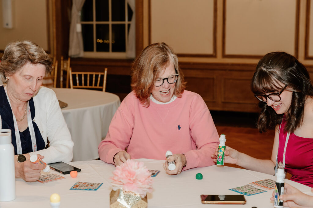 Three people are sitting at a round table playing bingo, using markers, and smiling in a warmly lit room with wood-paneled walls.