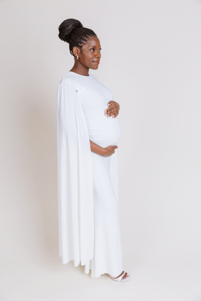 A person in a long white dress, standing sideways, cradling their pregnant belly against a plain light background.