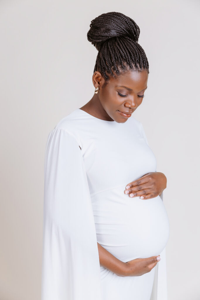 A person in a white dress gently holds their pregnant belly, standing against a plain, light-colored background.