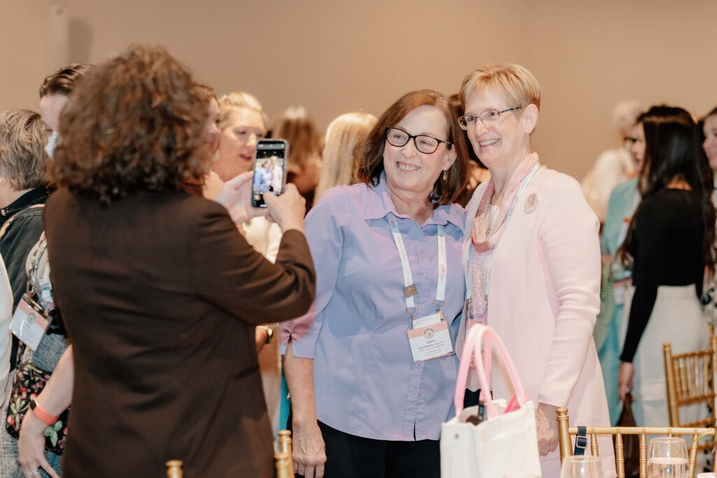 A person takes a photo of two people smiling at a social gathering, with others mingling in the background.