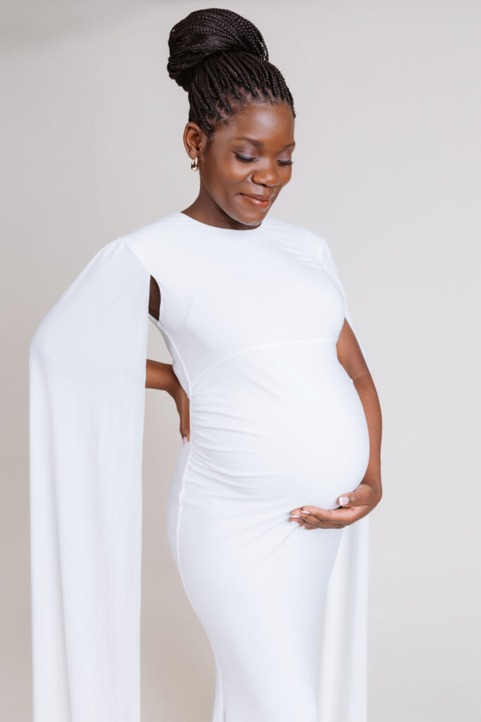 A person wearing a long, white dress gently holds their pregnant belly, smiling softly against a plain background.
