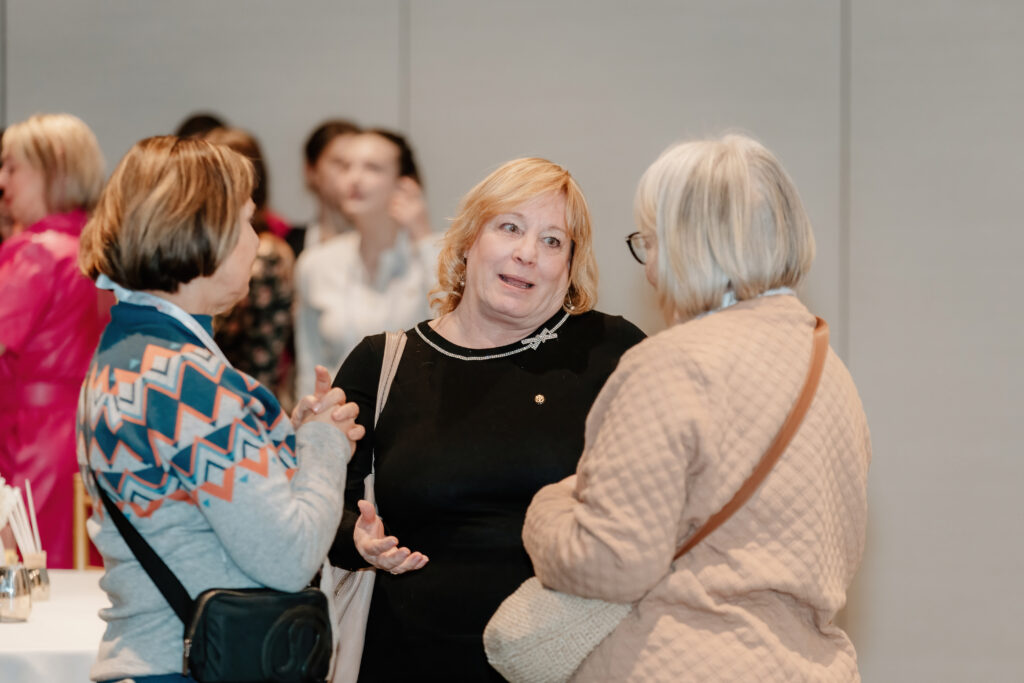 Three people engaged in conversation at an indoor event, with others in the background. Casual attire, friendly expressions, plain backdrop.