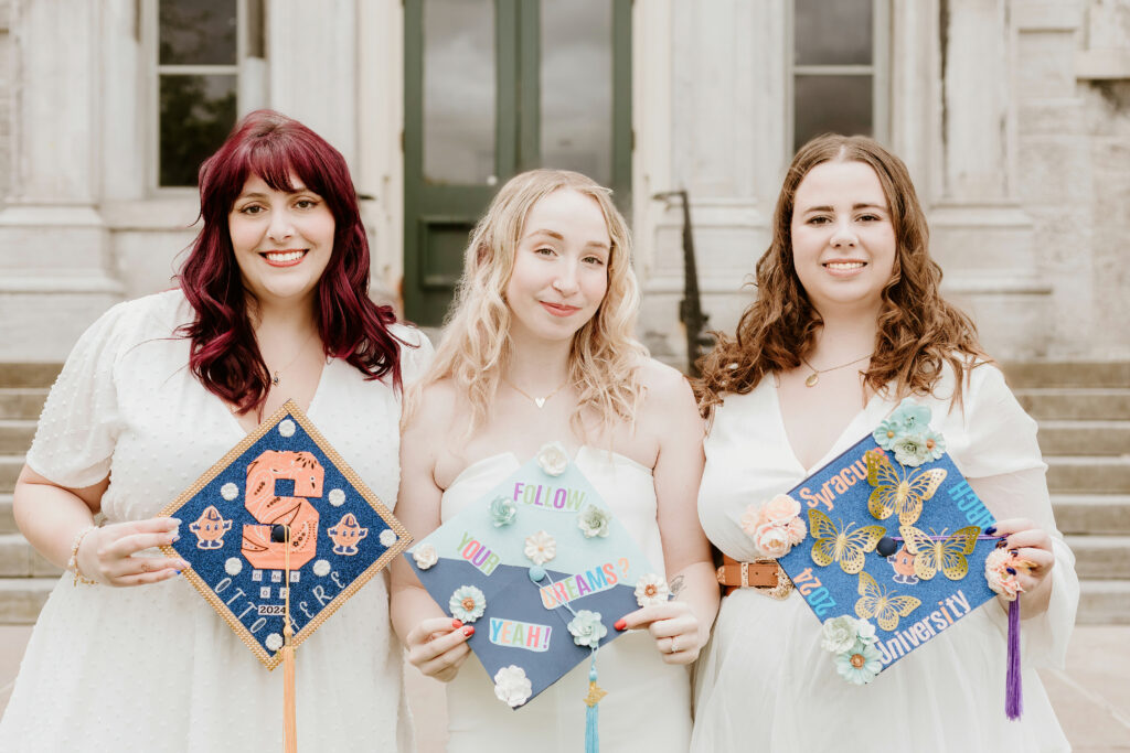 Three people are holding elaborately decorated graduation caps, smiling in front of a building, possibly celebrating their graduation or a similar achievement.