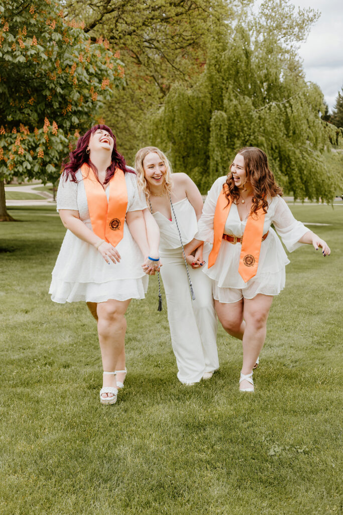 Three joyful people walk across a grassy field, wearing white outfits with orange graduation stoles and medals, laughing and holding hands.