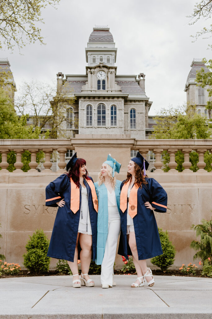 Three people in graduation attire smiling and standing in front of a historic building with a clock tower. They appear to be celebrating their achievement.