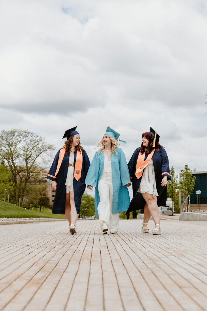 Three individuals in graduation gowns and caps walk joyfully together on a paved area, with cloudy skies and greenery in the background.