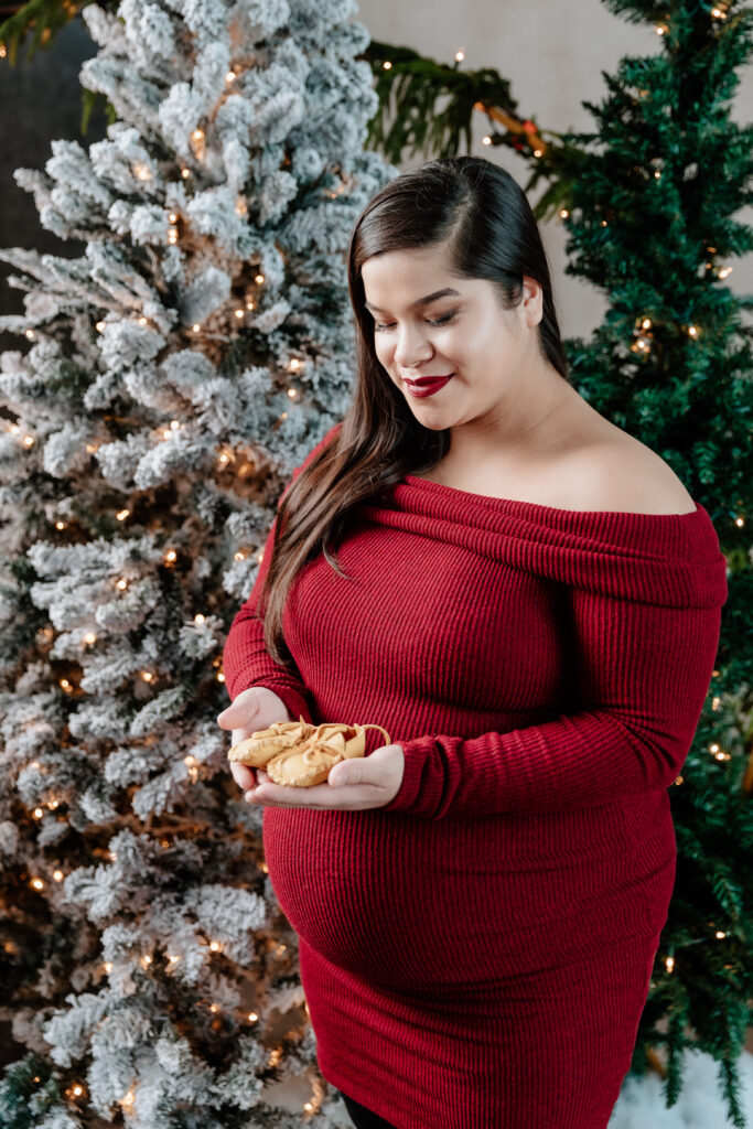 A person in a red sweater holds cookies, standing beside lit, snow-covered Christmas trees in a festive setting.