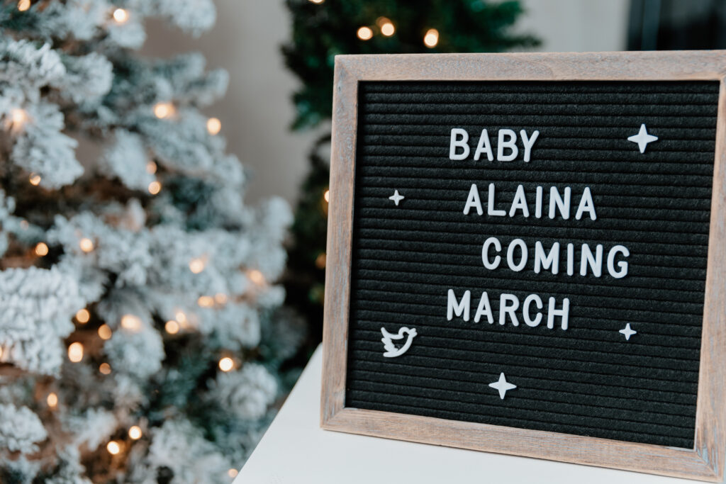 A letter board announces “Baby Alaina Coming March” beside a decorated, snow-covered Christmas tree with glowing lights in the background.