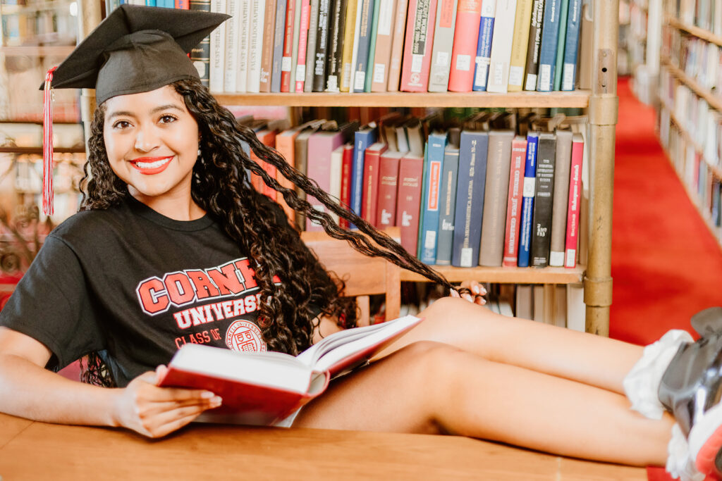 A person in a graduation cap sits among bookshelves, smiling while holding a red book, wearing a university T-shirt labeled