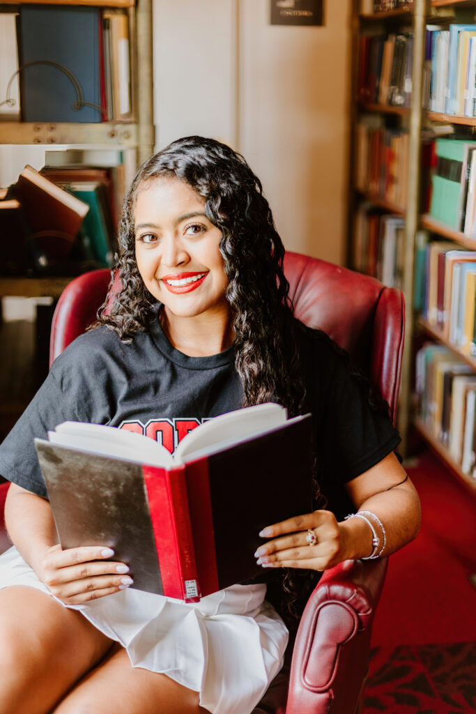 A person is seated in a red leather chair in a library, smiling while holding an open book, wearing a casual top and a skirt.