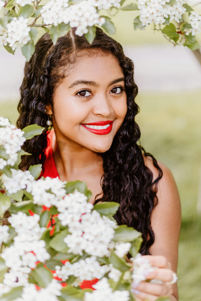 A person with curly hair smiling among white blossoms, wearing red lipstick and a red outfit, creating a cheerful springtime atmosphere.