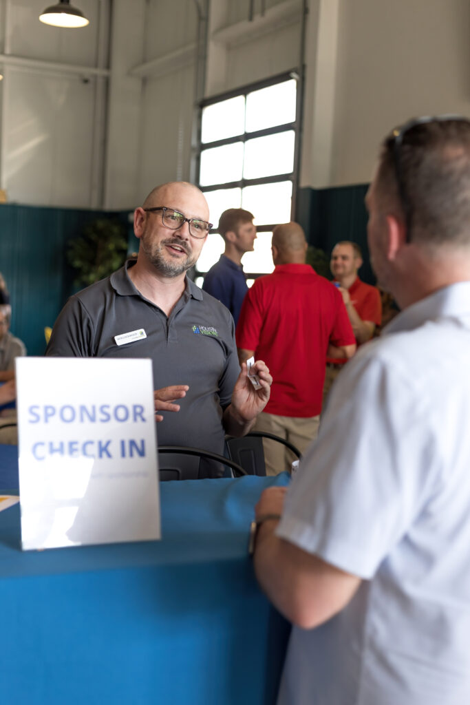 A person at a sponsor check-in table engages with another person at an indoor event; others converse in the background.