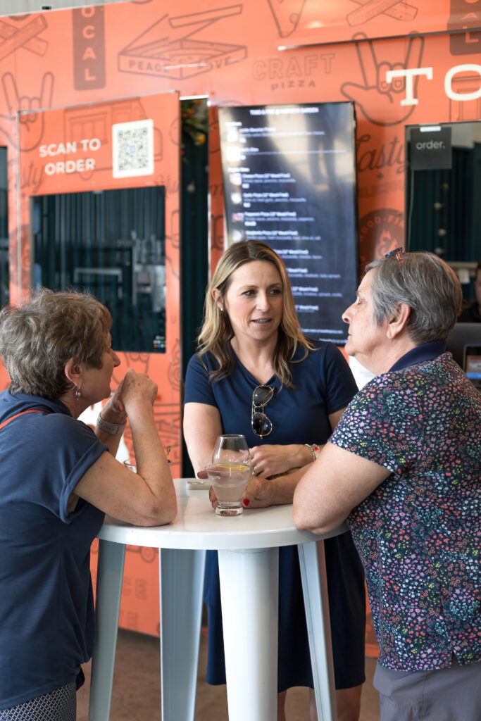 Three people converse at a table with drinks. A QR code and digital menu are visible on the orange wall behind them.