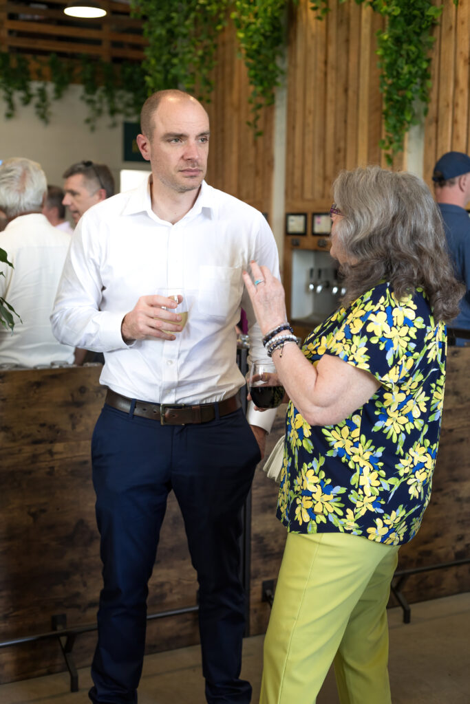 Two people in conversation at a social event, holding drinks. Indoor setting with wooden decor and hanging plants. Multiple people in background.