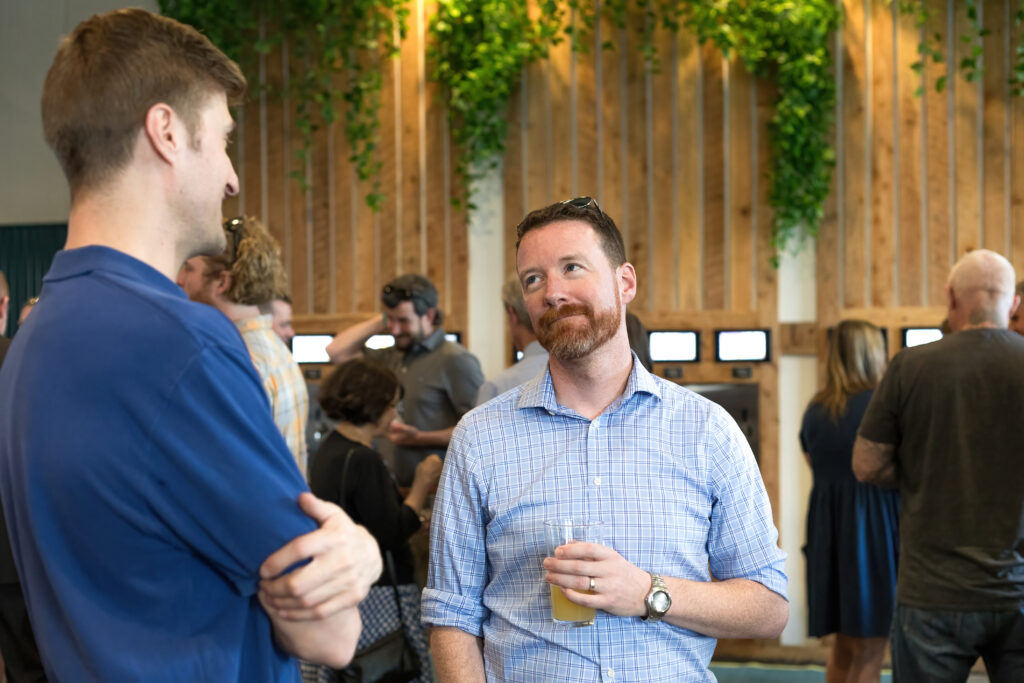 Two people conversing at an indoor gathering. Others mingle in the background. Walls decorated with plants and wooden panels. Casual, social atmosphere.
