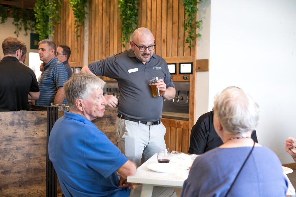 A person stands talking to seated individuals with drinks, in a room with wooden decor and green plants hanging from the ceiling.