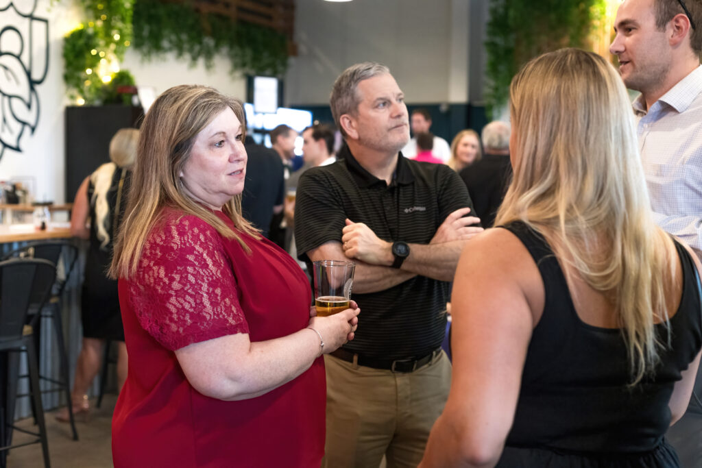 A group of people are conversing in a modern indoor setting, surrounded by greenery and casual decor, suggesting a social or networking event.