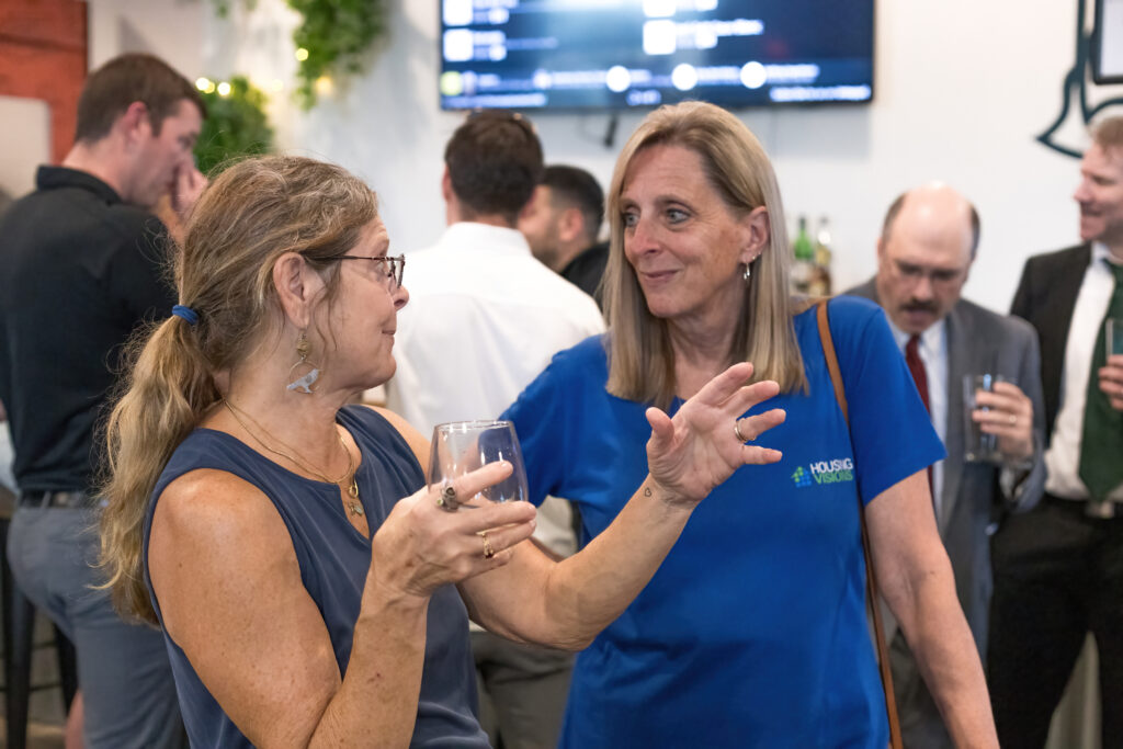 Two people are conversing at a social event, holding drinks. Others are in the background. Indoor setting with a screen displaying information.