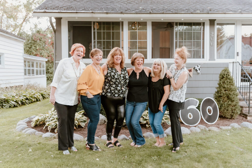 Six people smiling in front of a house, celebrating a 60th birthday, with large "60" numbers displayed on the grass.