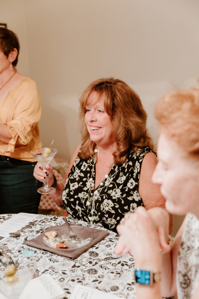 Three people are at a table sharing drinks and appetizers in a cozy room. One person holds a martini glass, smiling.