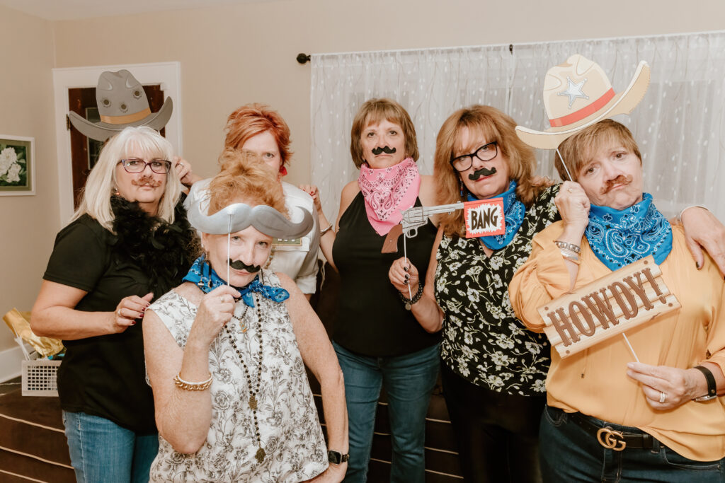 Six people with cowboy-themed props and mustaches pose playfully indoors, holding signs and wearing hats and bandanas.