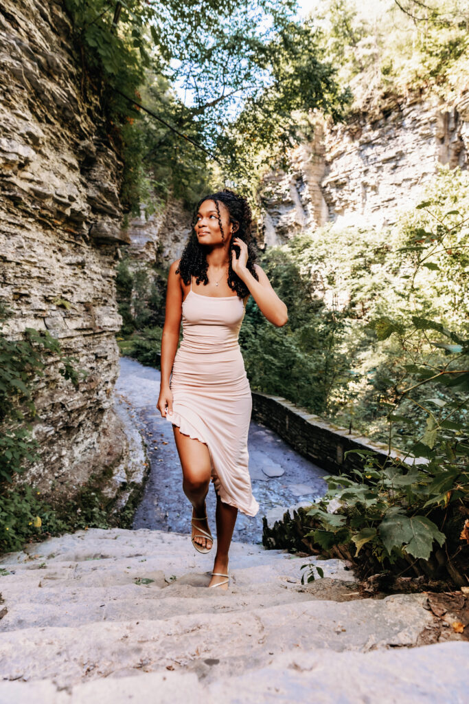 A person in a beige dress ascends a rocky, narrow path surrounded by lush greenery and tall cliffs in a serene natural setting.